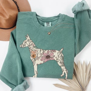May include: A teal green sweatshirt with a floral print of a dog. The dog is outlined in white and the flowers are in shades of pink, yellow, and orange. The sweatshirt has a crew neck and long sleeves.