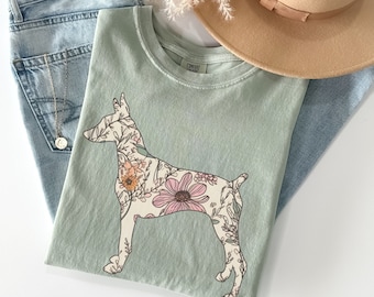 Può includere: Una maglietta verde chiaro con un design floreale di un cane. Il cane è delineato in nero e riempito con fiori rosa, arancioni e verdi.