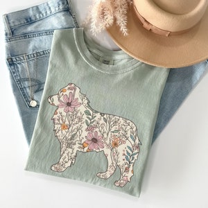May include: A light green t-shirt with a floral design in the shape of a dog. The dog is outlined in white and the flowers are in shades of pink, yellow, and orange.