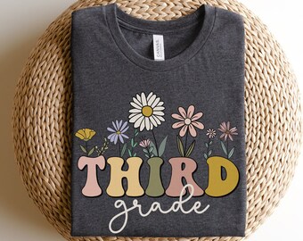 Op de afbeelding: Donkergrijs t-shirt met een bloemenontwerp en de woorden "THIRD grade" in een retro lettertype. De afbeelding toont kleurrijke bloemen in vazen. Het shirt is gemaakt van een zacht materiaal en wordt getoond op een geweven oppervlak.