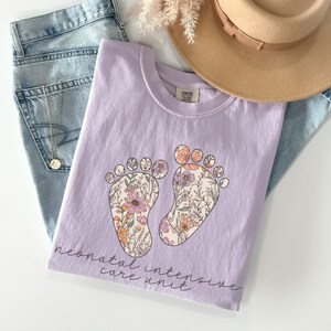 Puede incluir: Una camiseta lavanda con un estampado floral de dos huellas y el texto "neonatal intensive care unit".