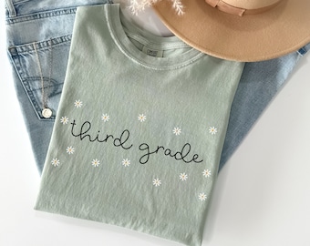 May include: A light green t-shirt with the text "third grade" in black script. The shirt is decorated with small white daisy flowers.