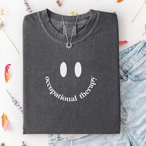 May include: A dark gray t-shirt with a smiley face design and the words "occupational therapy" in white. The shirt is paired with denim shorts and a silver necklace. The word "Pepper" is in the bottom left corner.