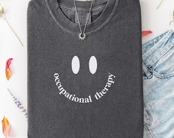May include: A dark gray t-shirt with a smiley face design and the words "occupational therapy" in white. The shirt is paired with denim shorts and a silver necklace. The word "Pepper" is in the bottom left corner.