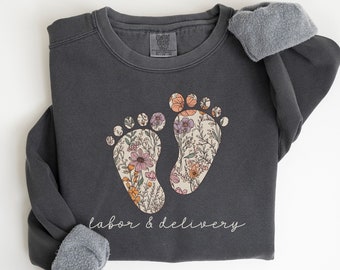 May include: A dark gray sweatshirt with a floral design of two baby footprints and the text "labor & delivery".