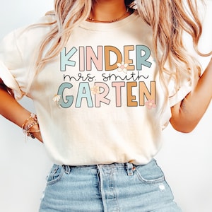 Peut inclure: Un t-shirt blanc avec le texte "Kinder mrs. smith garten" en lettres colorées avec des fleurs. Les lettres sont soulignées en noir et les fleurs sont en rose, jaune et bleu.