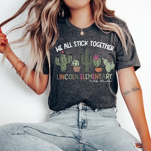 May include: A grey t-shirt with the text "We all stick together" and a row of cactus illustrations. Below the text is "Lincoln Elementary" and "Mrs. Smith".
