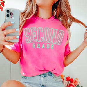 Puede incluir: Una camiseta rosa brillante con las palabras "SECOND GRADE" en letras grandes de color verde ne&oacute;n. La camiseta se combina con pantalones cortos de mezclilla. La imagen est&aacute; bien iluminada y tiene una est&eacute;tica suave y ligeramente borrosa.