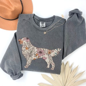 May include: A gray sweatshirt with a floral print of a golden retriever. The sweatshirt has a crew neck and long sleeves. The print is in shades of pink, yellow, and green.