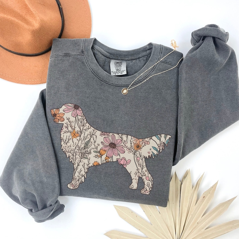 May include: A gray sweatshirt with a floral print of a golden retriever. The sweatshirt has a crew neck and long sleeves. The print is in shades of pink, yellow, and green.