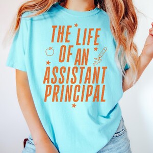 Può includere: T-shirt azzurro con la scritta arancione "THE LIFE OF AN ASSISTANT PRINCIPAL". Il design include una mela, una matita e delle stelle.