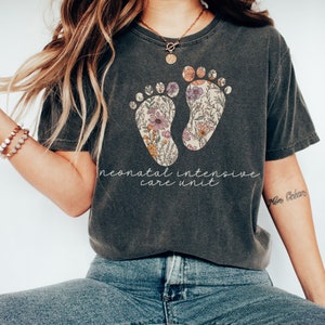 Puede incluir: Una camiseta gris oscuro con un dise&ntilde;o floral de dos huellas y el texto "neonatal intensive care unit".