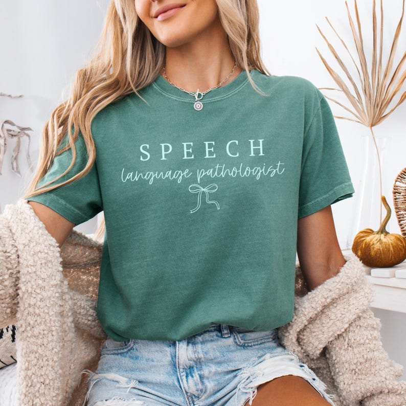 K&ouml;nnte beinhalten: T&uuml;rkisfarbenes T-Shirt mit wei&szlig;em Aufdruck &bdquo;SPEECH language pathologist&ldquo; und einer Schleife. Das Shirt hat einen Rundhalsausschnitt und kurze &Auml;rmel.