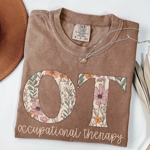 K&ouml;nnte beinhalten: Ein braunes T-Shirt mit den Buchstaben "OT" in einem floralen Design. Die Buchstaben sind von Blumen und Bl&auml;ttern in Rosa-, Orange- und Gr&uuml;nt&ouml;nen umgeben. Der Text "occupational therapy" steht unter den Buchstaben.