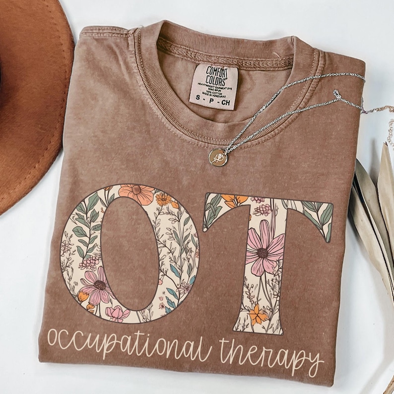 K&ouml;nnte beinhalten: Ein braunes T-Shirt mit den Buchstaben "OT" in einem floralen Design. Die Buchstaben sind von Blumen und Bl&auml;ttern in Rosa-, Orange- und Gr&uuml;nt&ouml;nen umgeben. Der Text "occupational therapy" steht unter den Buchstaben.