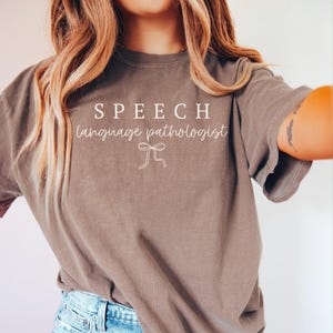 K&ouml;nnte beinhalten: Ein braunes T-Shirt mit den Worten "SPEECH language pathologist" in wei&szlig;er Schrift. Eine wei&szlig;e Schleifen-Grafik befindet sich unter dem Text. Das Shirt hat einen l&auml;ssigen Stil.