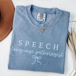 K&ouml;nnte beinhalten: Ein blaues Comfort Colors T-Shirt mit dem wei&szlig;en Schriftzug "SPEECH language pathologist". Eine goldene Halskette mit Anh&auml;nger liegt auf dem Shirt. Ein brauner Hut ist oben links zu sehen.
