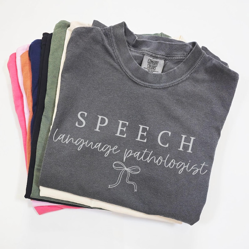 K&ouml;nnte beinhalten: Ein Stapel gefalteter T-Shirts in verschiedenen Farben, darunter Rosa, Orange, Marineblau, Schwarz, Gr&uuml;n und Creme. Das oberste Shirt ist anthrazitfarben und tr&auml;gt die Aufschrift "SPEECH language pathologist" und eine Schleifen-Grafik.