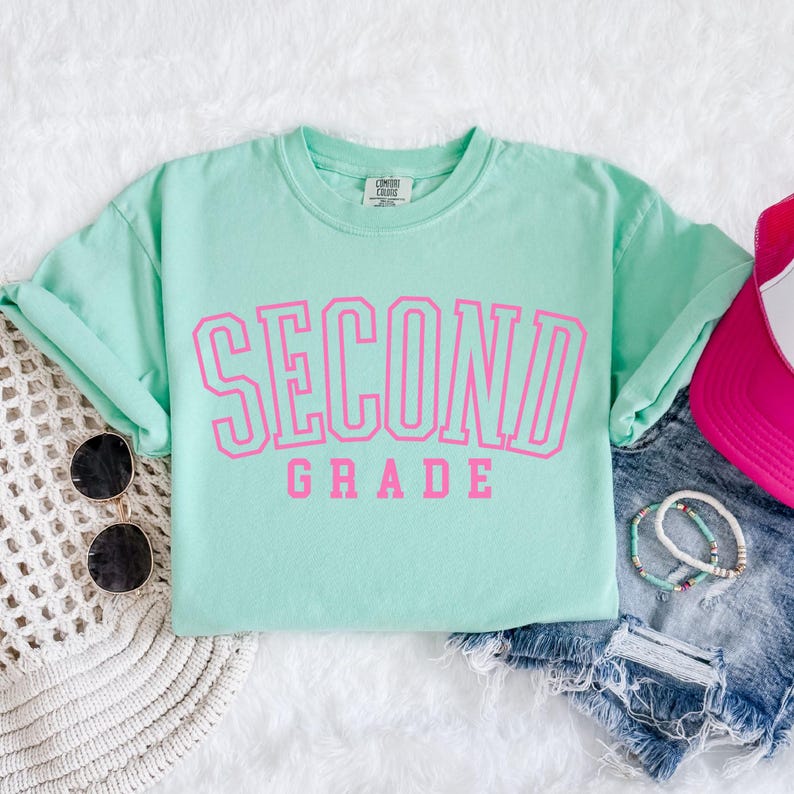 Puede incluir: Una camiseta verde menta con las palabras "SECOND GRADE" en letras rosas brillantes. La camiseta est&aacute; combinada con gafas de sol, una gorra rosa y pantalones cortos de mezclilla. La camiseta es de un material suave.
