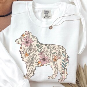 Pu&ograve; includere: Felpa bianca con una silhouette di pastore australiano con motivo floreale. Il cane &egrave; delineato in nero e riempito con una variet&agrave; di fiori rosa, arancioni e verdi.