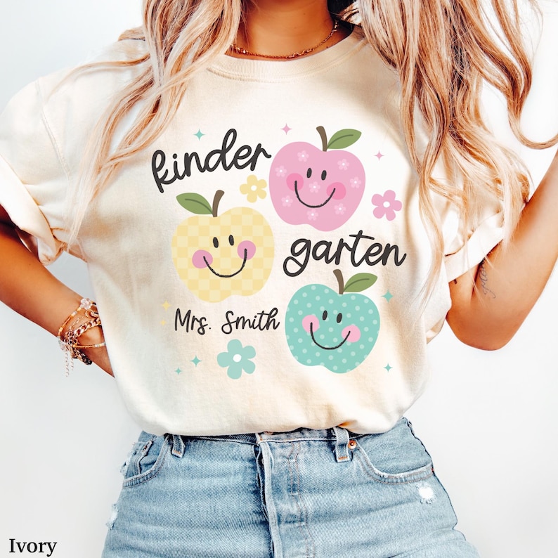 May include: Ivory-colored t-shirt with the words "Kindergarten" and "Mrs. Smith" printed on it. The design includes smiling apple graphics in pink, yellow, and teal, along with small flower and star accents.