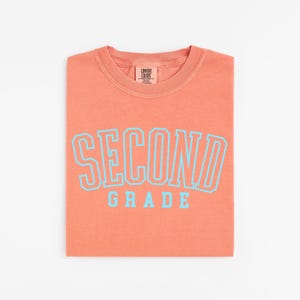Puede incluir: Una camiseta coral doblada con las palabras "SECOND GRADE" en letras grandes de color azul claro. La camiseta tiene cuello redondo y una peque&ntilde;a etiqueta en la parte superior. La camiseta est&aacute; hecha de un material suave.