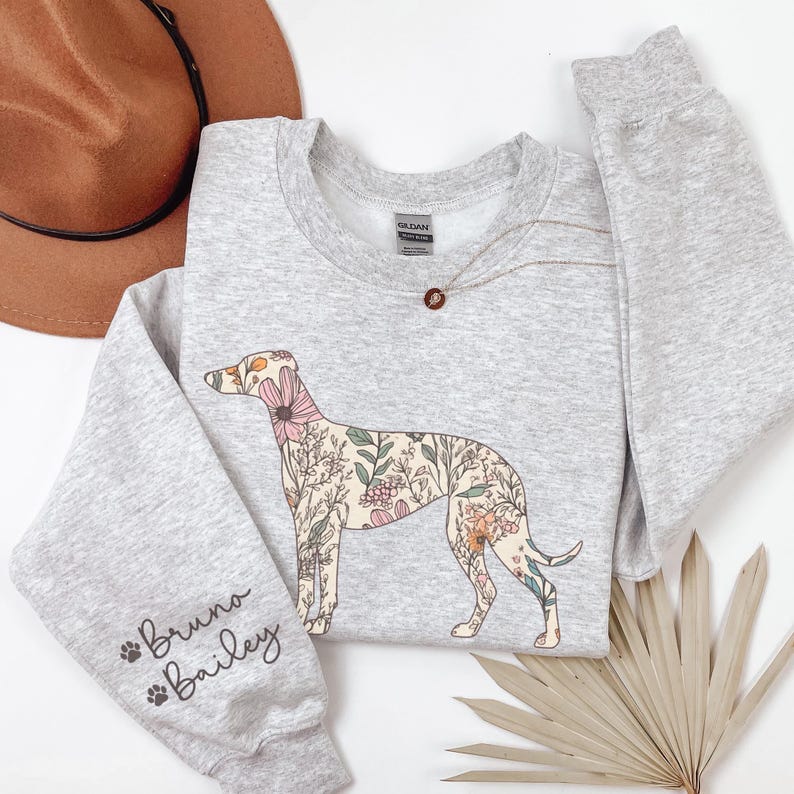 Peut inclure: Un sweat-shirt gris clair avec un motif de chien floral. Le chien est rempli de fleurs et de feuilles roses, beiges et vertes. La manche gauche porte le texte "Bruno Bailey". Un chapeau marron et un collier sont &eacute;galement visibles.