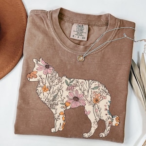 Könnte beinhalten: Ein braunes T-Shirt mit einem floralen Design einer Hundesilhouette. Das Shirt hat ein Etikett mit der Aufschrift "Comfort Colors" und "S - P - CH".