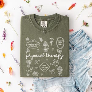 Puede incluir: Camiseta verde salvia con las palabras "physical therapy" e ilustraciones blancas. La camiseta se muestra con pantalones cortos de mezclilla y un collar plateado. Flores secas y p&eacute;talos est&aacute;n dispuestos alrededor de la ropa.