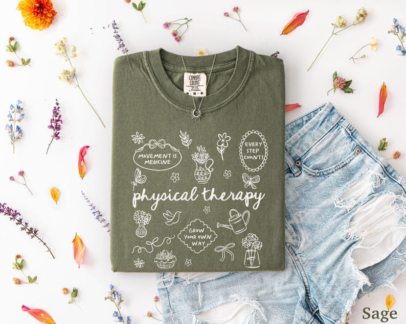 Puede incluir: Camiseta verde salvia con las palabras "physical therapy" e ilustraciones blancas. La camiseta se muestra con pantalones cortos de mezclilla y un collar plateado. Flores secas y p&eacute;talos est&aacute;n dispuestos alrededor de la ropa.
