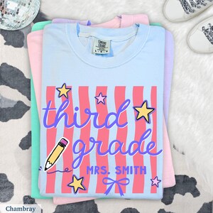 May include: A stack of light blue, pink, and mint green t-shirts. The top shirt is light blue with a graphic that says "third grade Mrs. Smith" in purple script with pink stripes, stars, a pencil, and a bow.