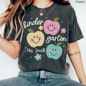 May include: A dark gray t-shirt with the word "kindergarten" and the name "Mrs. Smith" printed on it. The shirt features three smiling apple graphics in yellow, pink, and teal, with floral accents. The shirt is made of a soft material.