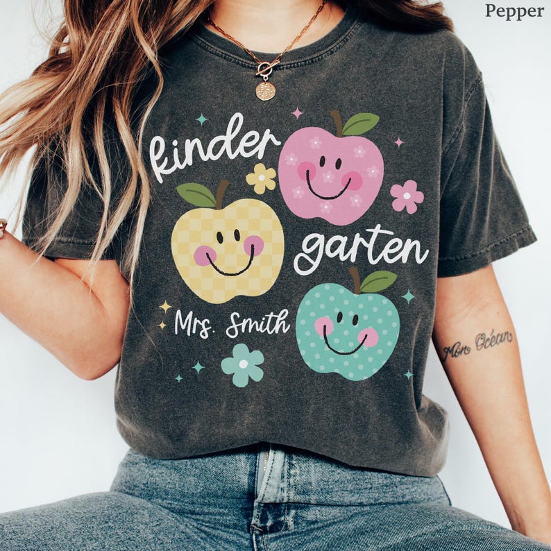 May include: A dark gray t-shirt with the word "kindergarten" and the name "Mrs. Smith" printed on it. The shirt features three smiling apple graphics in yellow, pink, and teal, with floral accents. The shirt is made of a soft material.