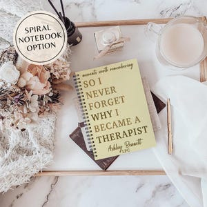 May include: A spiral notebook with a pale yellow cover featuring the text 'moments worth remembering SO I NEVER FORGET WHY I BECAME A THERAPIST Ashley Bennett LPC' in dark brown lettering, accompanied by dried flowers, a glass mug, and a pen.