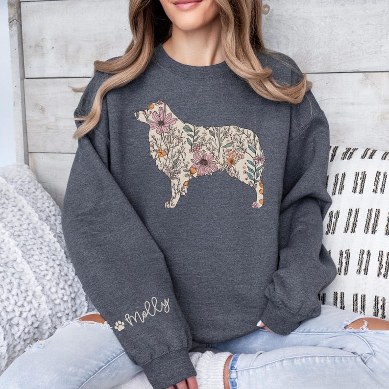 Puede incluir: Una sudadera gris oscuro con una silueta de perro pastor australiano con estampado floral. El perro est&aacute; contorneado en blanco y el estampado floral es en tonos de rosa, amarillo y verde. La palabra "Molly" est&aacute; bordada en la manga izquierda en blanco con una huella de pata.
