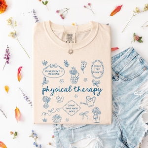 Puede incluir: Camiseta color marfil con la frase "physical therapy" e ilustraciones azules. La camiseta se combina con pantalones cortos de mezclilla y un collar plateado. Acentos florales y p&eacute;talos est&aacute;n esparcidos alrededor de la camiseta.