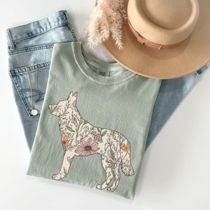 May include: A light green t-shirt with a floral design in the shape of a German Shepherd dog. The design features pink, yellow, and orange flowers.