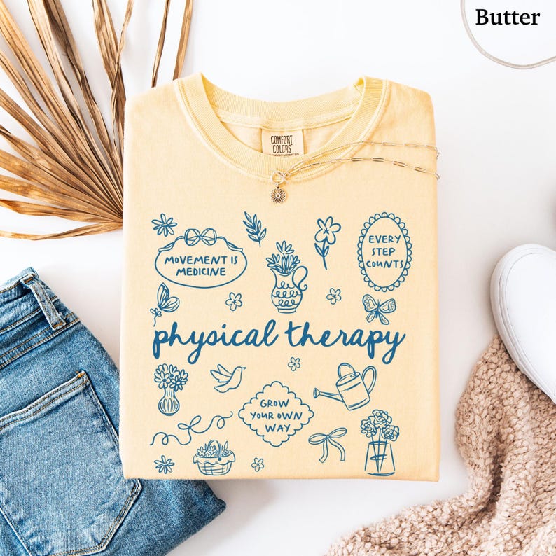 Puede incluir: Una sudadera de color mantequilla con la frase "physical therapy" en azul. La camisa presenta varias ilustraciones en azul, incluyendo flores, mariposas y las frases "Movement is Medicine" y "Every Step Counts."