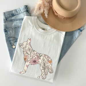 May include: White t-shirt with a floral design in the shape of a dog. The flowers are in shades of pink, orange, and green.