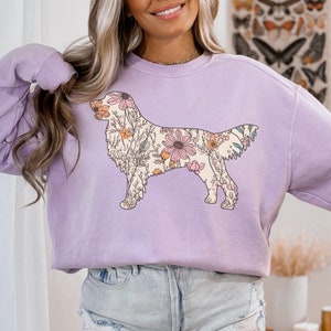 May include: A lavender sweatshirt with a floral design of a golden retriever. The floral design is in shades of pink, orange, and yellow.