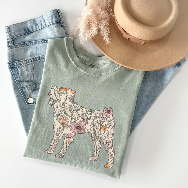 May include: A light green t-shirt with a floral pug silhouette design. The pug is outlined in black and filled with a variety of pink, orange, and yellow flowers.