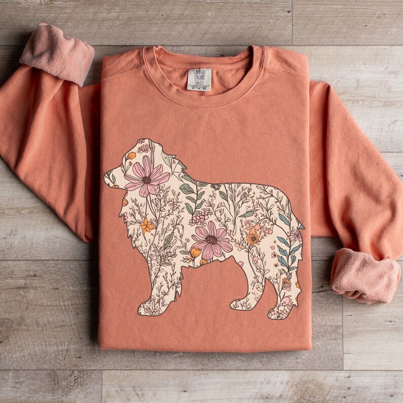 Pu&ograve; includere: Una felpa color pesca con un disegno floreale a forma di cane. I fiori sono rosa, bianchi e gialli.