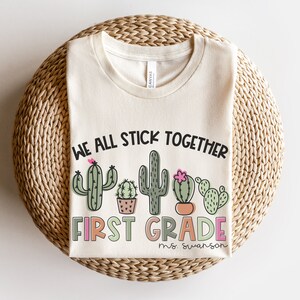 May include: A beige t-shirt with the text "We all stick together first grade" and a row of cactus illustrations. The text "ms. swanson" is below the cactus illustrations.