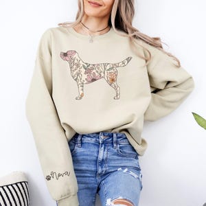 May include: A light beige sweatshirt with a floral Labrador Retriever design. The sweatshirt has a crew neck and long sleeves. The text "Nova" is printed on the left sleeve.