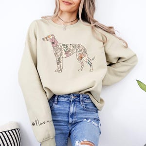 Peut inclure: Un sweat-shirt beige avec un motif de chien floral. Le chien est rempli de diverses fleurs roses, p&ecirc;che et vertes. Le nom "Nova" est imprim&eacute; sur la manche en &eacute;criture cursive avec une empreinte de patte. Le sweat-shirt est de coupe ample.