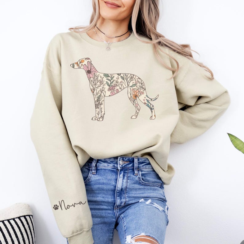 Peut inclure: Un sweat-shirt beige avec un motif de chien floral. Le chien est rempli de diverses fleurs roses, p&ecirc;che et vertes. Le nom "Nova" est imprim&eacute; sur la manche en &eacute;criture cursive avec une empreinte de patte. Le sweat-shirt est de coupe ample.