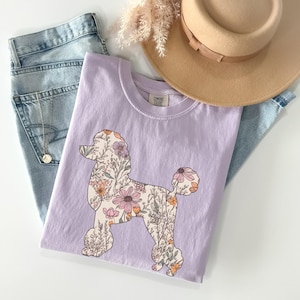 May include: A lavender t-shirt with a floral poodle silhouette design. The text "COMFORT COLORS" is printed on the shirt.