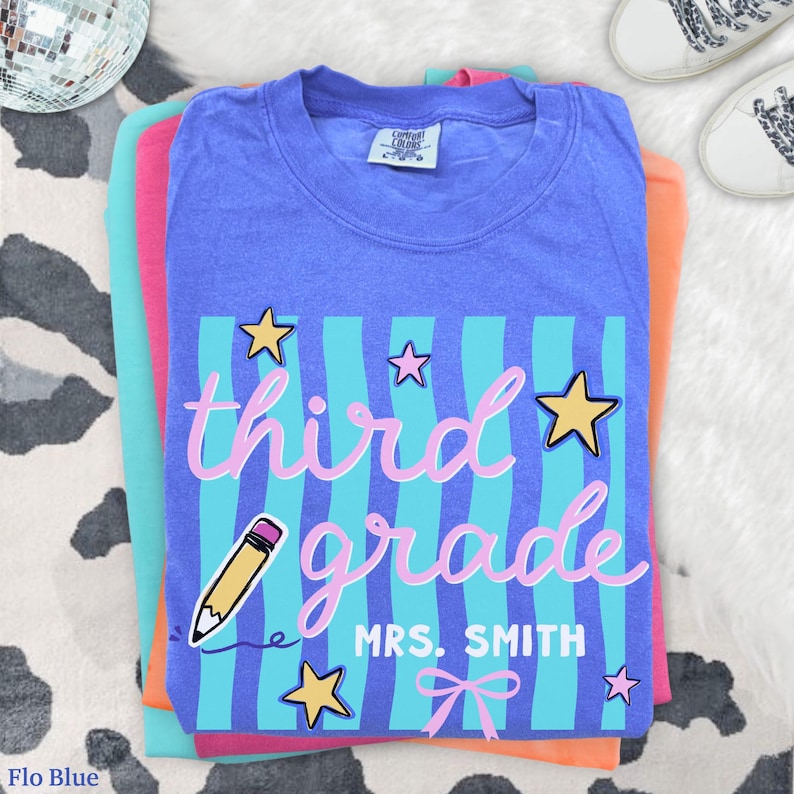 May include: A stack of colorful t-shirts, with a blue shirt on top featuring the words "third grade" in pink script, with a pencil and stars. The shirt also says "MRS. SMITH".