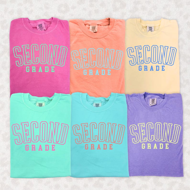 Puede incluir: Seis camisetas coloridas en rosa, naranja, amarillo, turquesa, azul claro y morado. Cada camiseta tiene las palabras "SECOND GRADE" impresas en la parte delantera en un color contrastante. Las camisetas est&aacute;n dobladas y expuestas sobre un fondo claro.