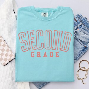 Puede incluir: Una camiseta azul claro con las palabras "SECOND GRADE" en letras color coral. La camiseta est&aacute; doblada junto a unos vaqueros azules, un su&eacute;ter blanco, una funda de tel&eacute;fono y una pulsera dorada. Marca Comfort Colors.